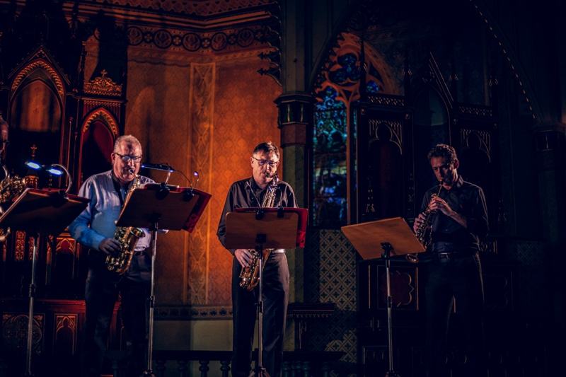 Três saxofonistas tocando diante de um altar iluminado e vitrais